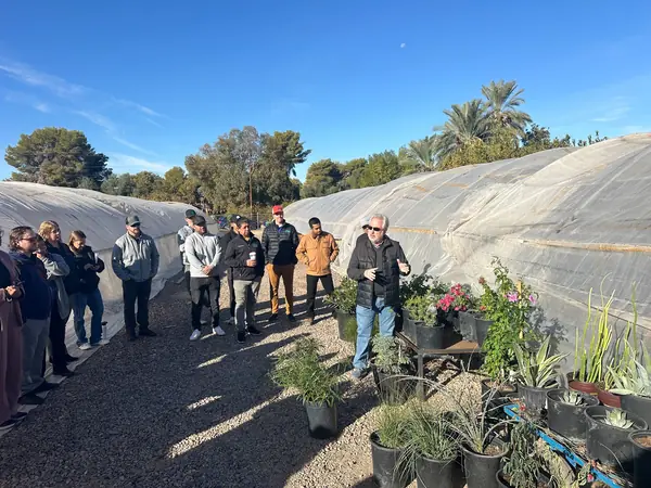 Group touring rows of nursery plants beside shade-cloth greenhouses during nursery training