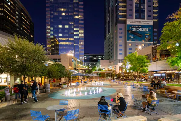 CityScape plaza at night with shade trees, lit fountains and people at outdoor tables beneath downtown towers