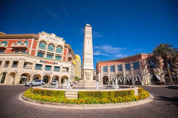 Tivoli Village plaza in Las Vegas with its obelisk fountain ringed by yellow and red flower beds, palms and arcaded buildings