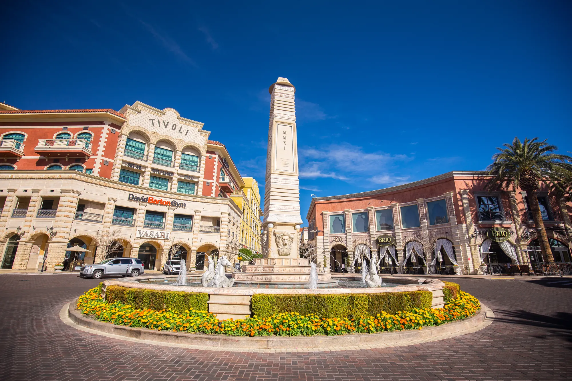 Tivoli Village plaza in Las Vegas with its obelisk fountain ringed by yellow and red flower beds, palms and arcaded buildings